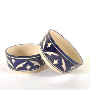 May include: Two small ceramic bowls with a cream-colored interior and a deep blue exterior. The outer surface features a repeating white hand-painted pattern of stylized leaf and star shapes, with thin white lines bordering the top and bottom rims.