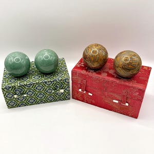 May include: Two pairs of Chinese health balls, one pair green and the other brown with gold accents, rest on decorative boxes. The green balls sit on a green and blue patterned box, while the brown balls are on a red box.