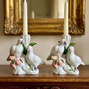 May include: A pair of white ceramic candle holders, each featuring two sculpted birds perched on a base with pink fruit and green leaves. A tall, slender cream-colored taper candle is inserted into the central holder of each piece.
