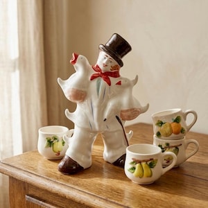 Puede incluir: Una figura de cerámica de una persona con sombrero de copa y pañuelo rojo, sosteniendo dos gansos blancos, acompañada de tres tazas pequeñas de cerámica blanca decoradas con ilustraciones de frutas en relieve, incluyendo peras y naranjas.