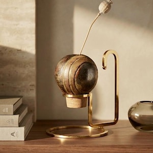 May include: A decorative object featuring a spherical, multi-toned brown and metallic orb resting on a light-coloured cork base. This assembly is supported by a gold-toned metal stand with a circular base and a curved arm holding a dried poppy pod.