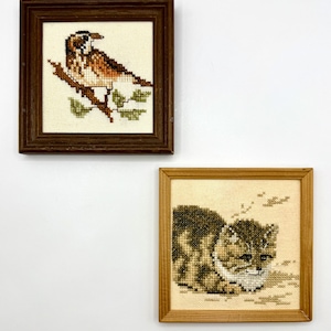 May include: Two square framed cross-stitch embroideries. The top piece features a brown and white bird perched on a branch in a dark wood frame. The bottom piece shows a brown tabby cat in a light wood frame, both on a plain white background.