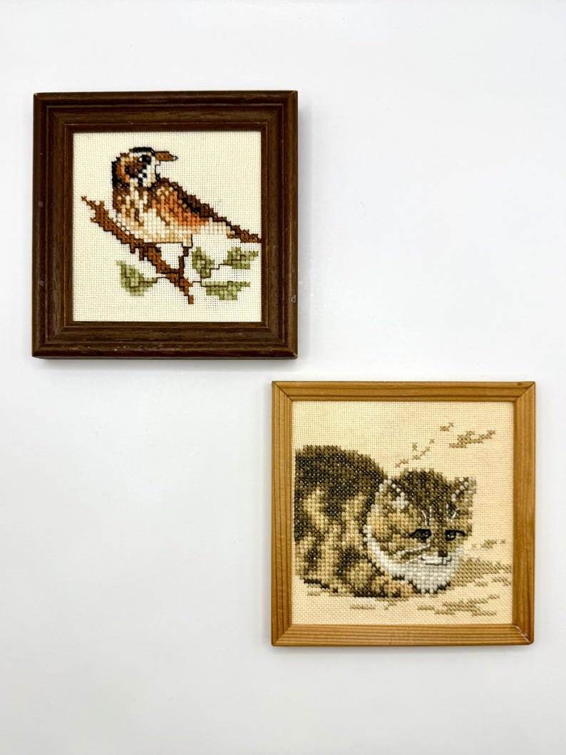 May include: Two square framed cross-stitch embroideries. The top piece features a brown and white bird perched on a branch in a dark wood frame. The bottom piece shows a brown tabby cat in a light wood frame, both on a plain white background.