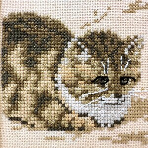 May include: A cross-stitch embroidery of a brown and tan tabby cat with dark stripes, white facial markings, and dark eyes. The cat is depicted in a seated position against a light cream-colored fabric background, framed in a light wood border.