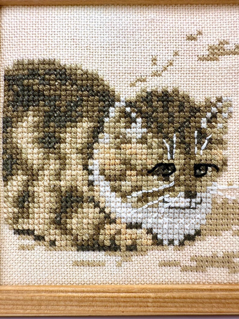 May include: A cross-stitch embroidery of a brown and tan tabby cat with dark stripes, white facial markings, and dark eyes. The cat is depicted in a seated position against a light cream-colored fabric background, framed in a light wood border.
