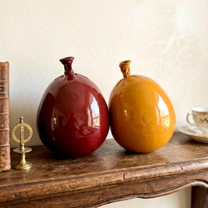 May include: Two ceramic vases, one deep red and the other golden yellow, sit side-by-side on a wooden shelf. The vases have a rounded, balloon-like shape with a small, textured neck. The shelf is made of dark wood.
