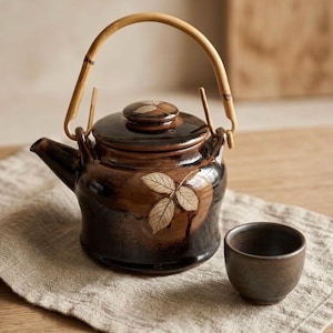 Peut inclure: Une théière en céramique marron avec une anse en bambou et une tasse assortie. La théière présente un motif de feuilles et repose sur un tissu en lin beige. L'ensemble à thé est conçu pour servir le thé.