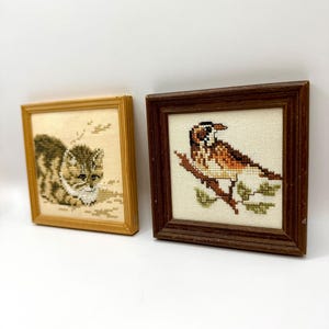 May include: Two square framed cross-stitch artworks. The left frame is light wood containing a stitched brown and white cat. The right frame is dark wood containing a stitched brown and white bird perched on a branch with green leaves.