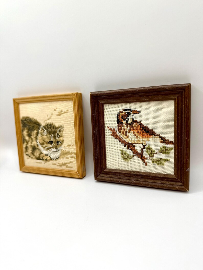 May include: Two square framed cross-stitch artworks. The left frame is light wood containing a stitched brown and white cat. The right frame is dark wood containing a stitched brown and white bird perched on a branch with green leaves.