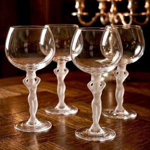 Puede incluir: Cuatro copas de vino transparentes con un diseño único. Cada copa presenta una figura esbelta y esmerilada como tallo, sosteniendo la copa. Las copas están dispuestas sobre una superficie de madera oscura, con un candelabro encendido al fondo.