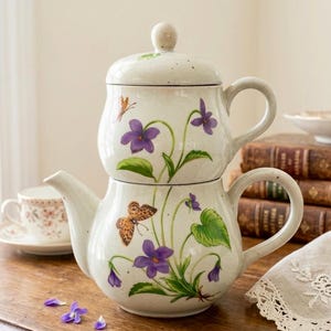 Pode incluir: Um conjunto de chá de cerâmica de duas peças. A chaleira e a tampa de cor creme são adornadas com violetas roxas, folhas verdes e borboletas laranja. A chaleira tem um bico curvo e uma alça arredondada. A tampa tem um pequeno botão.
