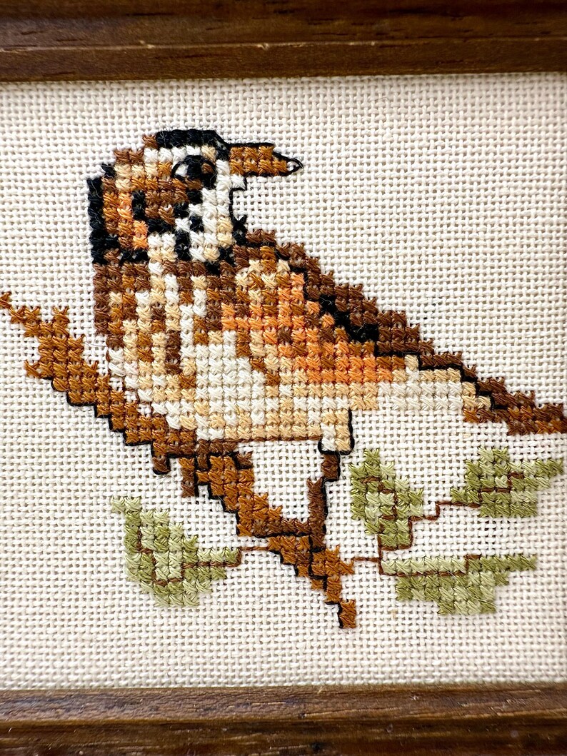 May include: A cross-stitch embroidery featuring a small brown and white bird perched on a brown branch with light green leaves. The bird has black markings on its head and wings, and the entire design is stitched onto a cream-colored fabric.