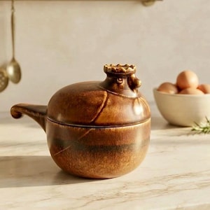 Może przedstawiać: Brązowy, szkliwiony ceramiczny pojemnik na jajka w kształcie kury ze zdejmowaną pokrywką. Pojemnik ma zaokrąglony korpus, rzeźbiony uchwyt w kształcie głowy kury na pokrywce oraz mały uchwyt boczny. Wykończenie w odcieniach ziemistego brązu.