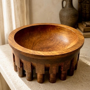 Peut inclure: Un bol rond en bois avec un intérieur lisse et une finition brun foncé. L'extérieur présente une série de supports verticaux en forme de blocs disposés en cercle autour de la base, créant une apparence surélevée et architecturale.