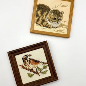 May include: Two square framed cross-stitch artworks. The top piece features a brown and white tabby cat in a light wood frame. The bottom piece displays a brown and white bird perched on a branch with green leaves, set in a dark wood frame.