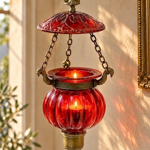 Può includere: Un portacandele sospeso con un contenitore in vetro rosso a coste, appeso tramite tre catene metalliche a una cupola in vetro rossa. La struttura presenta dettagli in metallo color ottone, inclusi una base a punta e ganci decorativi.