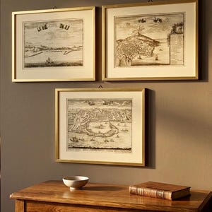 May include: Three framed antique maps with gold frames hang on a brown wall. The maps are black and white illustrations of coastal towns and landscapes. A wooden table with a bowl and a book sits below the maps.