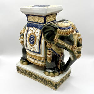 May include: A ceramic elephant garden stool with a square top and base. The elephant is primarily dark green with blue, white, and gold accents. The stool has intricate detailing and a decorative design.