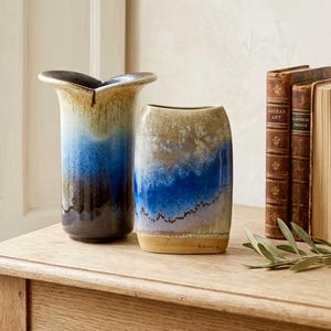 May include: Two ceramic vases with a blue, brown, and tan color scheme. The vase on the left has a flared top. The vase on the right is rectangular with a rounded top. The vases are on a wooden table.