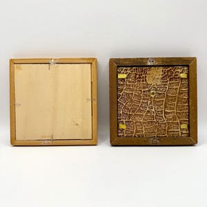 May include: Two square wooden frames shown from the back. The left frame has a light-colored, smooth wooden backing secured with metal staples. The right frame features a dark brown, textured backing with a crocodile-skin pattern and gold clips.