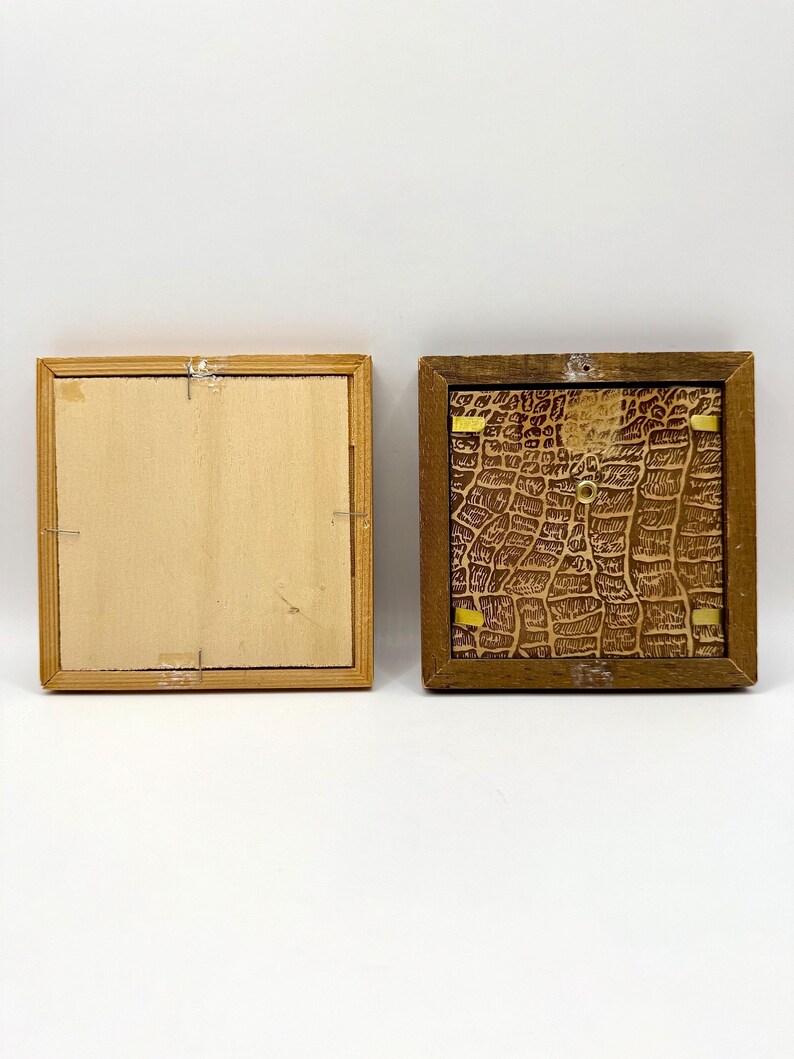 May include: Two square wooden frames shown from the back. The left frame has a light-colored, smooth wooden backing secured with metal staples. The right frame features a dark brown, textured backing with a crocodile-skin pattern and gold clips.