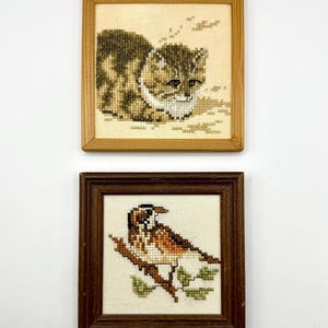 May include: Two square framed cross-stitch artworks. The top piece features a brown tabby cat in a light wood frame. The bottom piece displays a brown and white bird perched on a branch with green leaves, enclosed in a dark brown wood frame.