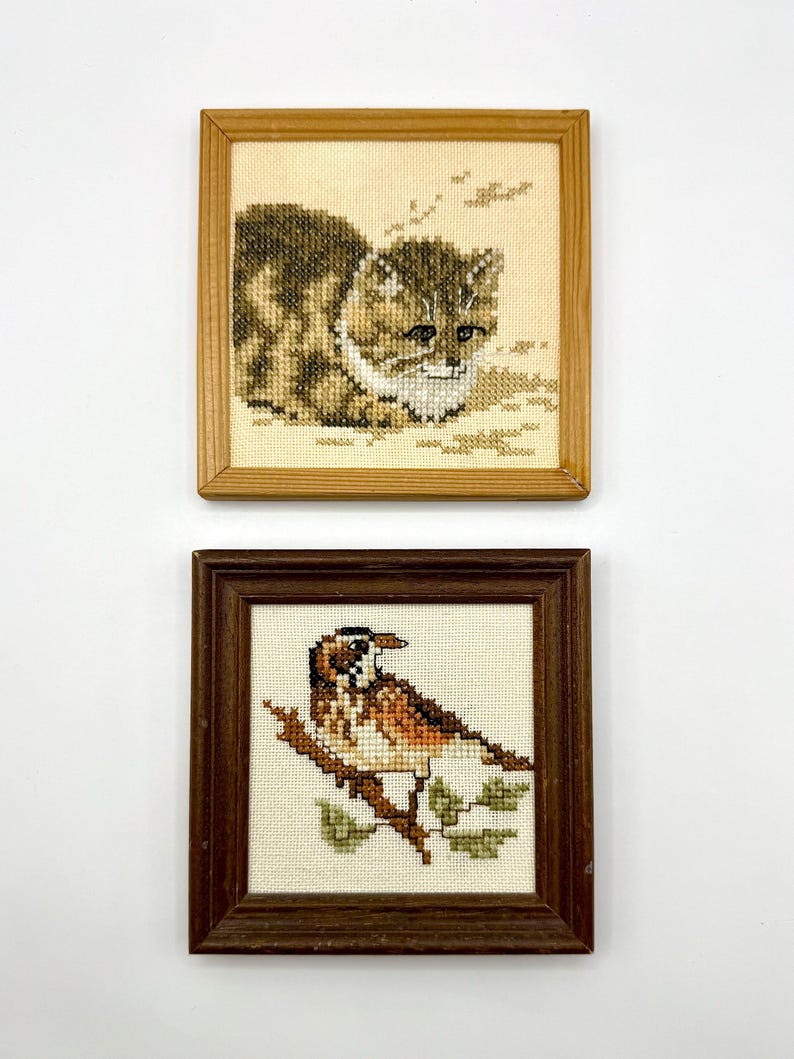 May include: Two square framed cross-stitch artworks. The top piece features a brown tabby cat in a light wood frame. The bottom piece displays a brown and white bird perched on a branch with green leaves, enclosed in a dark brown wood frame.
