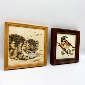 May include: Two square framed cross-stitch embroideries. The left piece features a brown and white tabby cat in a light wood frame. The right piece depicts a brown and white bird perched on a branch, enclosed in a dark brown wooden frame.