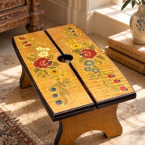 Puede incluir: Un reposapiés rectangular de madera con acabado marrón claro y un asa central recortada. La superficie superior presenta motivos florales pintados a mano en rojo, amarillo y azul, con hojas y tallos verdes que se extienden por ambos paneles.