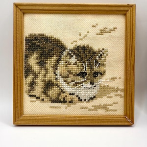 May include: A square wooden frame containing a cross-stitch embroidery of a brown and tan tabby kitten lying down. The kitten is depicted in shades of brown, tan, and white thread on a cream-colored fabric background with small stitched details.