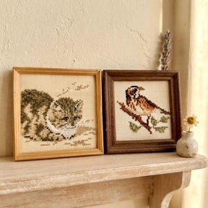 May include: Two square framed cross-stitch artworks. The left frame is light wood containing a stitched tabby cat. The right frame is dark wood containing a stitched bird perched on a branch. Both pieces are displayed on a wooden shelf.