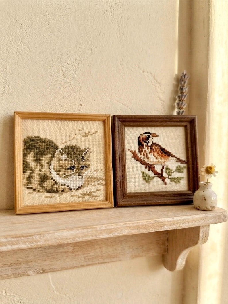 May include: Two square framed cross-stitch artworks. The left frame is light wood containing a stitched tabby cat. The right frame is dark wood containing a stitched bird perched on a branch. Both pieces are displayed on a wooden shelf.
