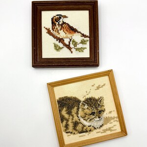 May include: Two square framed cross-stitch artworks. The top piece features a brown and white bird perched on a branch in a dark wood frame. The bottom piece displays a brown tabby cat with white markings in a light wood frame on cream fabric.