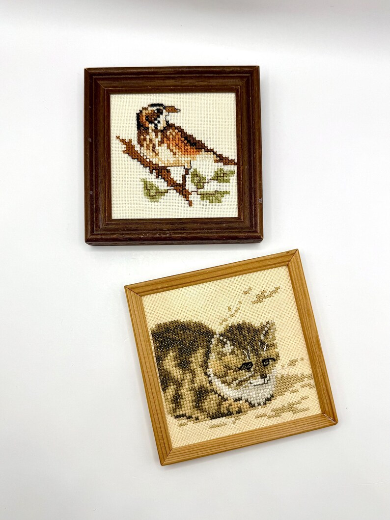May include: Two square framed cross-stitch artworks. The top piece features a brown and white bird perched on a branch in a dark wood frame. The bottom piece displays a brown tabby cat with white markings in a light wood frame on cream fabric.
