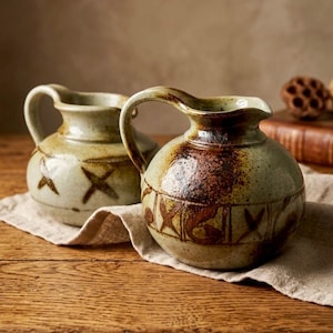 May include: Two handcrafted ceramic pitchers with loop handles. The pitchers have a speckled, earthy color palette of brown, tan, and green. Each pitcher features a decorative pattern of crosses and other shapes. They sit on a neutral-colored linen cloth.