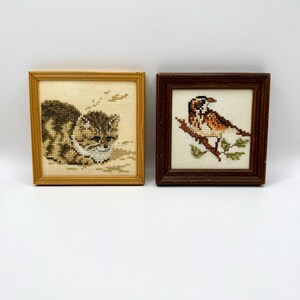 May include: Two square framed cross-stitch artworks. The left frame is light wood, containing a brown and white tabby cat. The right frame is dark wood, featuring a brown and white bird perched on a branch with green leaves on a cream background.