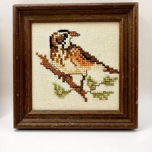 May include: A square cross-stitch embroidery featuring a brown and white bird perched on a branch with green leaves, set within a dark brown wooden frame. The bird has detailed plumage in shades of tan, brown, and black against a cream fabric.