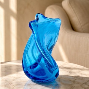 Pode incluir: Um vaso de vidro azul vibrante com uma forma escultural torcida e uma abertura larga e irregular no topo. O material translúcido exibe padrões de redemoinho e reflexos de luz por toda a sua estrutura orgânica e curva.