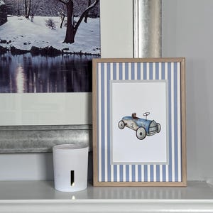 May include: A framed print with a watercolour illustration of a vintage blue race car, number 1. The frame has a blue and white striped border and a light wood frame. A white candle holder is to the left.
