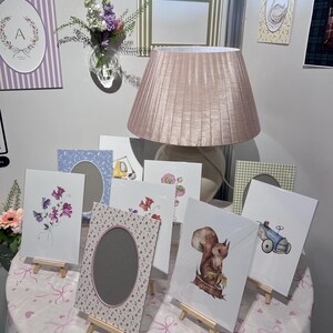 以下が含まれることがあります： 額装されたアートプリントと装飾フレームの展示。プリントには、リス、花、白鳥など、花や動物のイラストが描かれています。フレームは、楕円形や長方形など、さまざまなスタイルで、パステルカラーとパターンが使用されています。ピンク色のランプシェードがテーブルの上にあります。