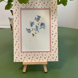 以下が含まれることがあります： 白い背景に青い花が描かれた、ピンクの縁取りのある額入りの花のプリント。 フレームは、小さな赤い花の模様が入ったクリーム色の背景です。 アートワークは小さな木製のイーゼルに展示されています。