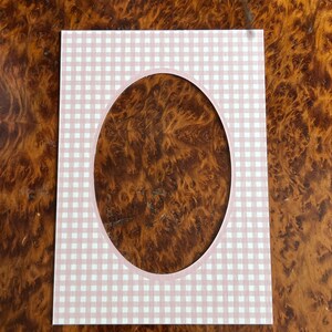 May include: A rectangular paper photo mat featuring a light pink and white gingham check pattern. The mat has a centered oval cutout with a thin, solid light pink border surrounding the opening, designed for framing photographs or artwork.