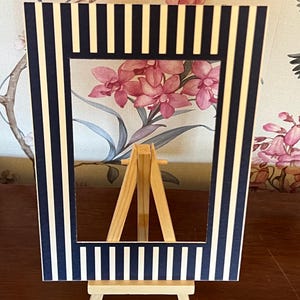Può includere: Una cornice rettangolare a righe blu navy e bianche, appoggiata su un piccolo cavalletto di legno. Lo sfondo presenta una carta da parati floreale con fiori rosa.