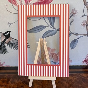 Può includere: Una cornice a righe rosse e bianche con un cavalletto in legno. La cornice ha un'apertura rettangolare ed è sostenuta da un piccolo cavalletto in legno naturale. Lo sfondo presenta un motivo di carta da parati floreale.