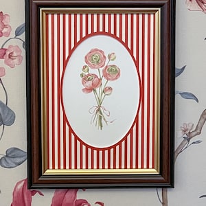 May include: Framed floral art print. The artwork features a bouquet of pink flowers tied with a ribbon, set against a red and white striped background. The frame is dark brown with a gold inner border, adding a classic touch to the piece.
