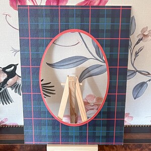 May include: A decorative picture frame with a blue, green, and pink plaid pattern, displayed on a small wooden easel. The frame has an oval aperture revealing a floral design. The background features floral wallpaper.