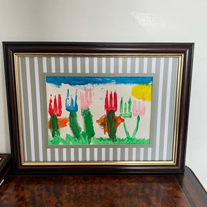 May include: A framed watercolor painting of colorful flowers and a yellow sun against a blue sky. The artwork is framed with a brown wooden frame, a gold inner frame, and a striped mat.