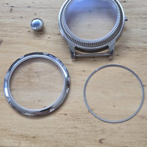 Pu&ograve; includere: Componenti dell'orologio, tra cui una cassa dell'orologio color argento con un cristallo trasparente, una lunetta, una corona e una guarnizione. La cassa dell'orologio ha una lunetta scanalata e anse per un cinturino. I componenti sono disposti su una superficie di legno.