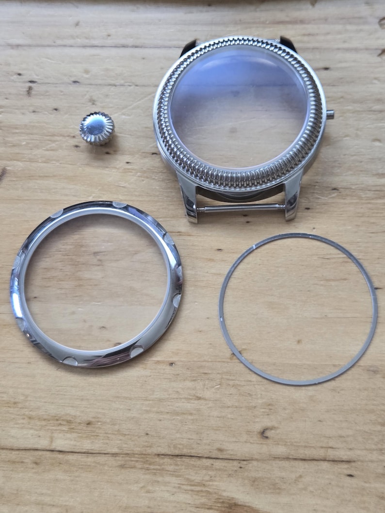 Pu&ograve; includere: Componenti dell'orologio, tra cui una cassa dell'orologio color argento con un cristallo trasparente, una lunetta, una corona e una guarnizione. La cassa dell'orologio ha una lunetta scanalata e anse per un cinturino. I componenti sono disposti su una superficie di legno.