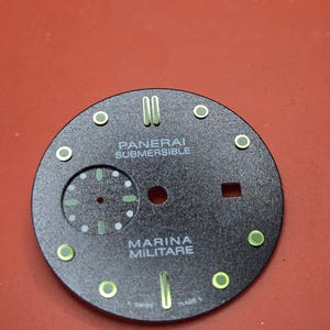 Pode incluir: Um mostrador de rel&oacute;gio preto com as palavras brancas &laquo;PANERAI SUBMERSIBLE&raquo; e &laquo;MARINA MILITARE&raquo;. Possui marcadores verdes e brancos, um pequeno mostrador de segundos e a inscri&ccedil;&atilde;o &laquo;SWISS MADE&raquo;.